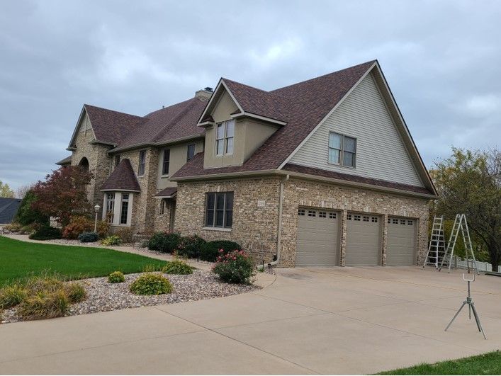 Large two-story house with brown roof, stone facade, tan garage doors, and a curved driveway on a cloudy day.