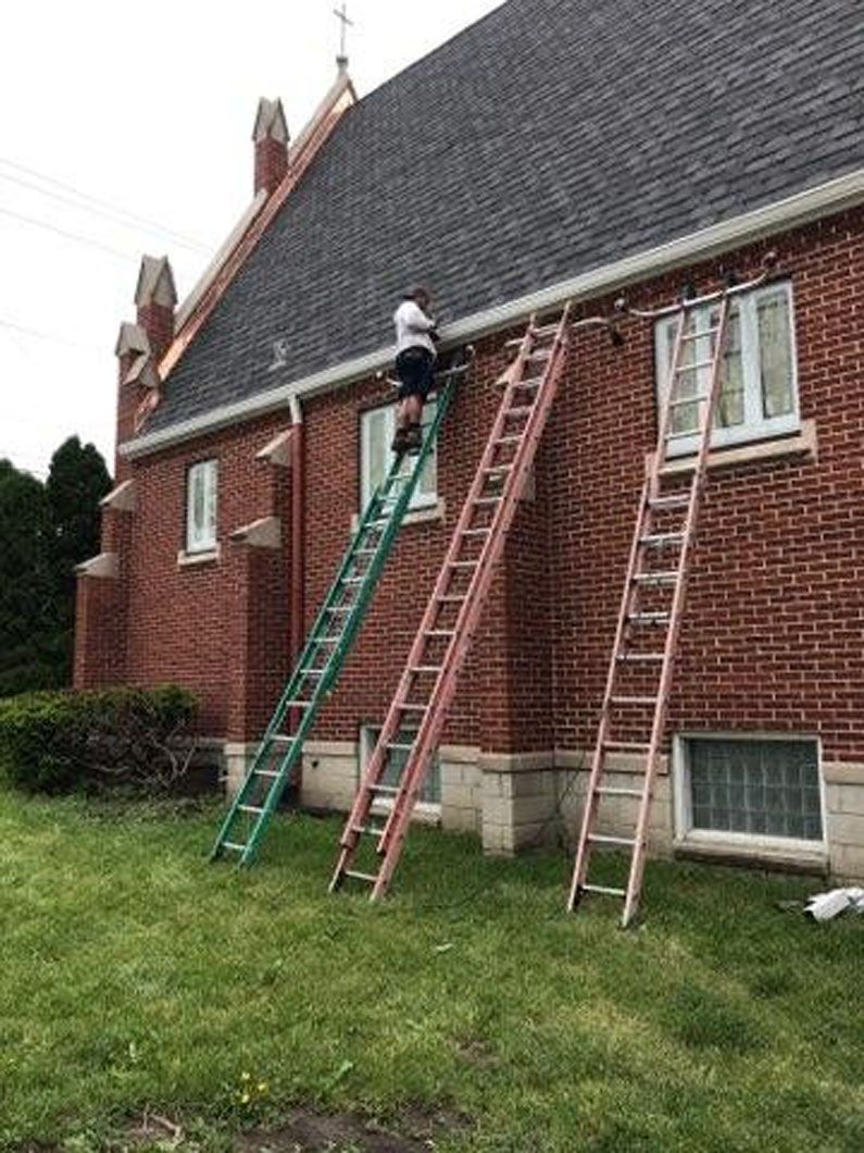 Person on ladders hanging lights on a red brick building with a dark roof. Three ladders are propped up.