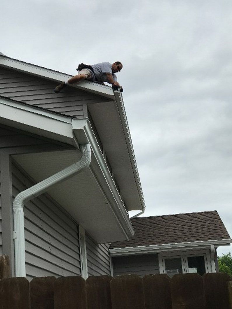 Man on a roof, cleaning a gutter. Overcast sky. Gray siding and brown shingles.