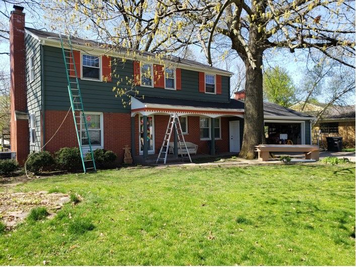 Two-story house with red brick and green siding; ladders lean against the house, indicating renovation work.