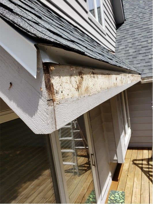 Damaged eaves on a two-story house, revealing rotted wood. Gray siding, white trim, and a deck visible.