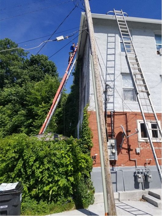 A utility worker on a ladder near power lines. The ladder leans against a pole next to a building.