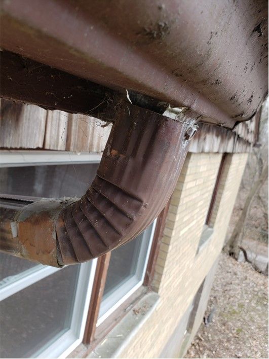 Damaged brown gutter downspout connected to a brown gutter on a brick building.