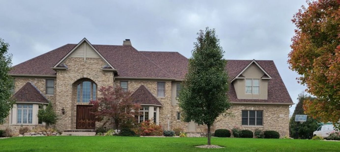 Large brick house with brown roof, green lawn, and trees under a cloudy sky.
