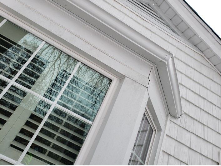 White-framed bay window on a shingled house reflects trees. The window frame has decorative trim.