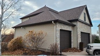 Two-story garage with stone and tan siding, brown roof, and two dark garage doors. A car is parked nearby.