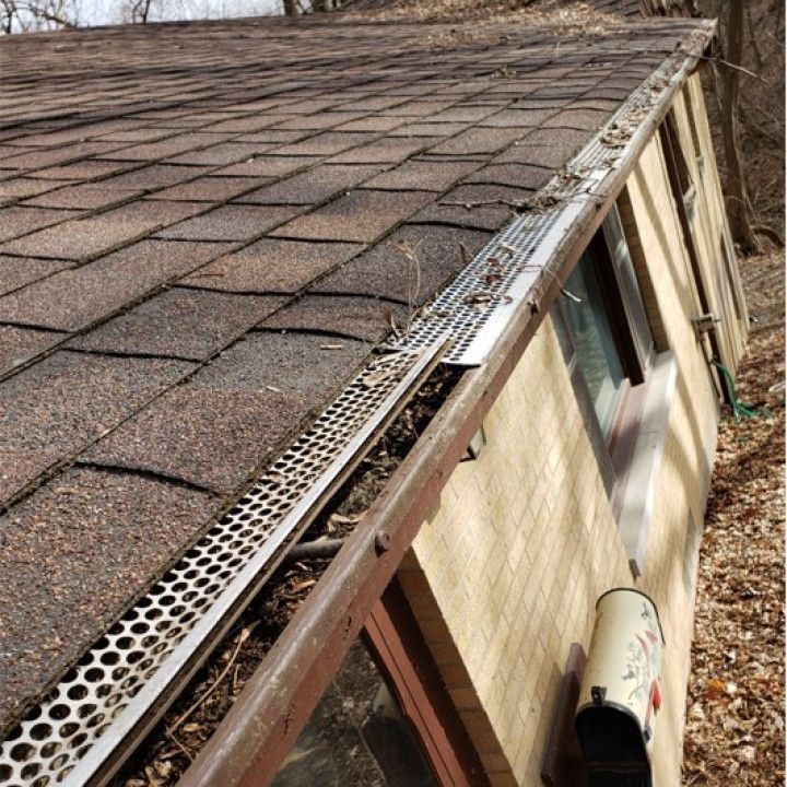 Gutter filled with debris on a brown shingled roof with a screen guard, next to a brick wall and a mailbox.