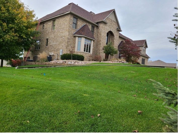 Large brick house on a grassy hill under a cloudy sky. Autumn colors are visible in the trees.