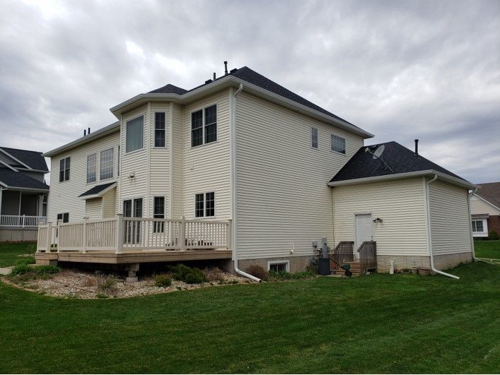 Two-story beige house with a deck and attached garage, under an overcast sky. Green lawn surrounds.