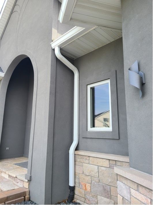 Gray stucco house with white gutters, a window, and a stone base.