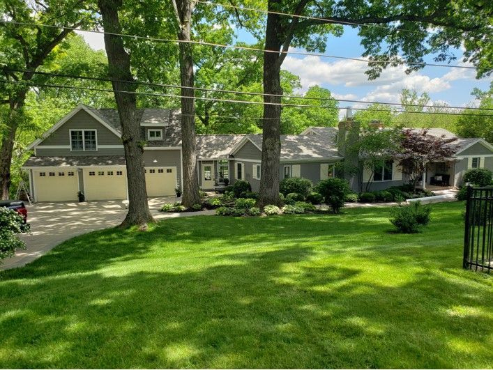 A large gray house with a two-car garage and lush green lawn, trees frame the view on a sunny day.