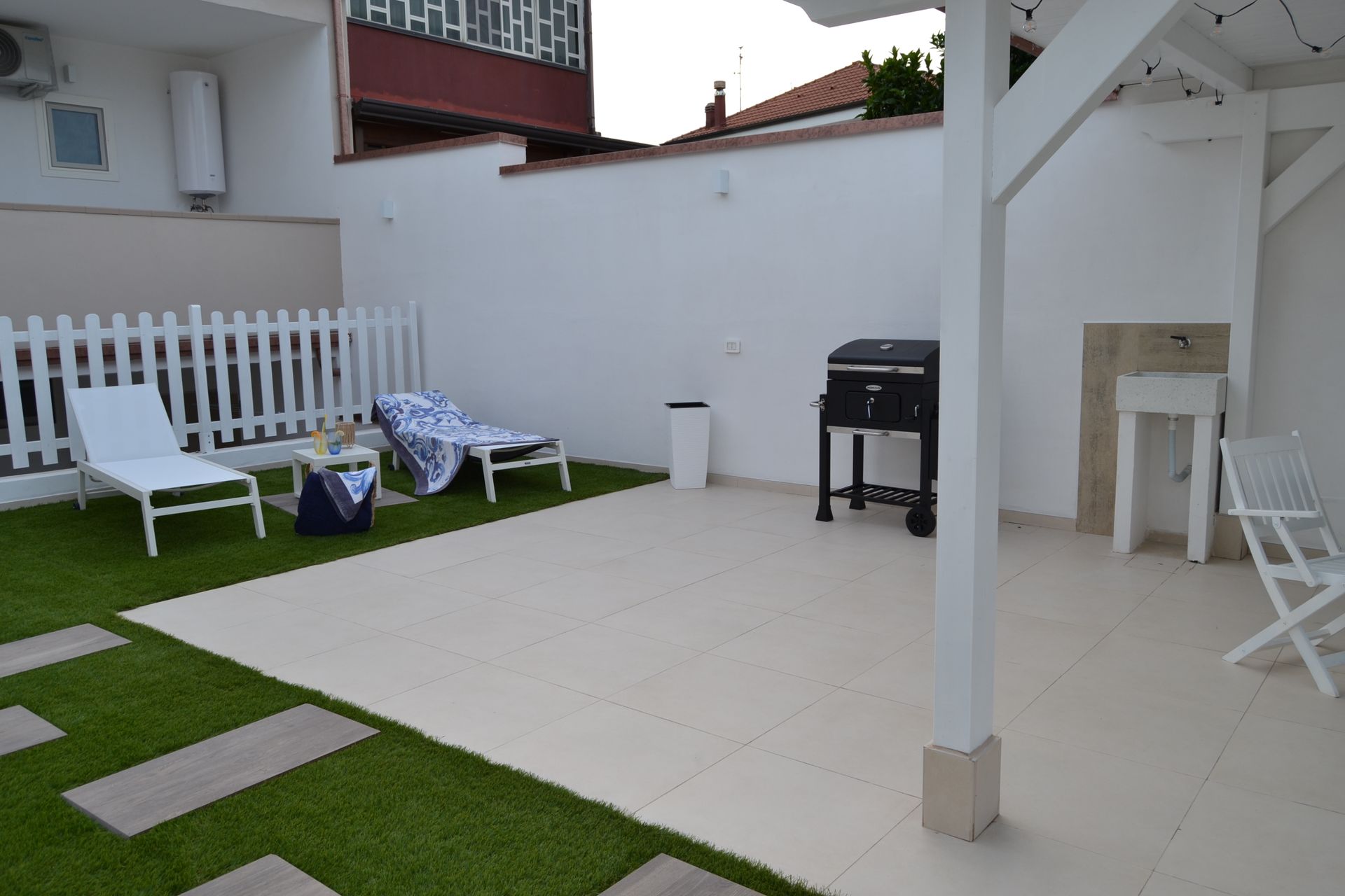 Patio con panchine bianche, prato verde, sdraio e barbecue accanto a un muro bianco.