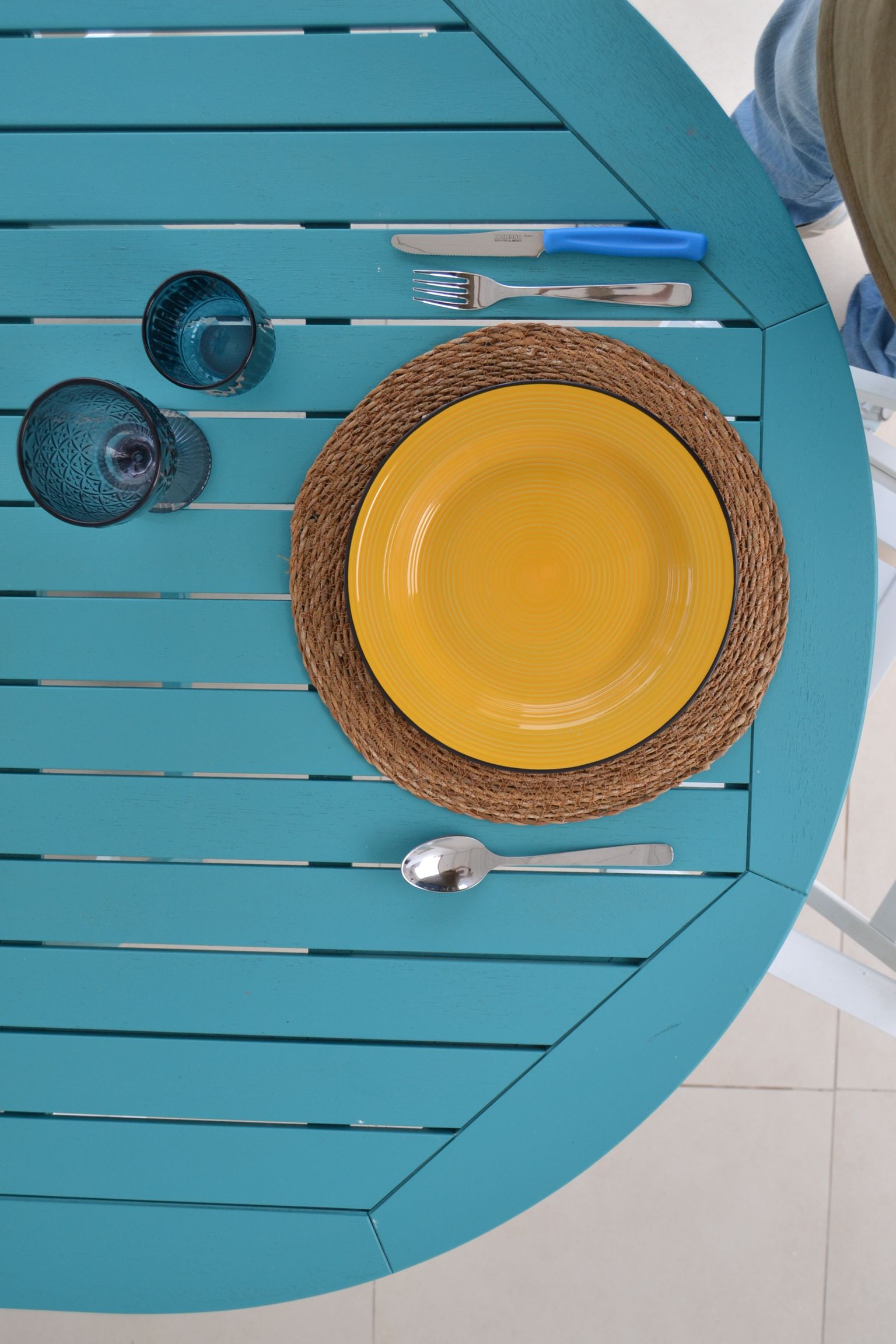 Servizio da tavola turchese con piatto giallo, sottopiatto intrecciato, bicchieri blu e posate.