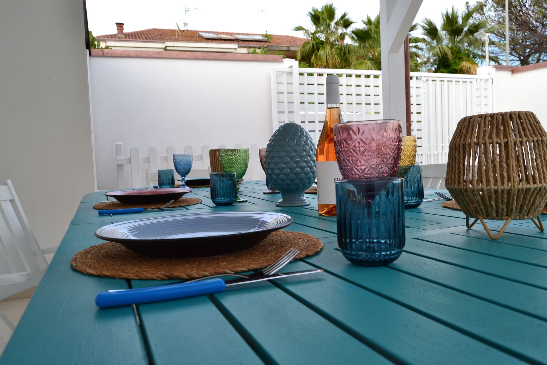 Tavolo da esterno blu con tovagliette intrecciate, tazze colorate e una lanterna a forma di cestino su un patio.