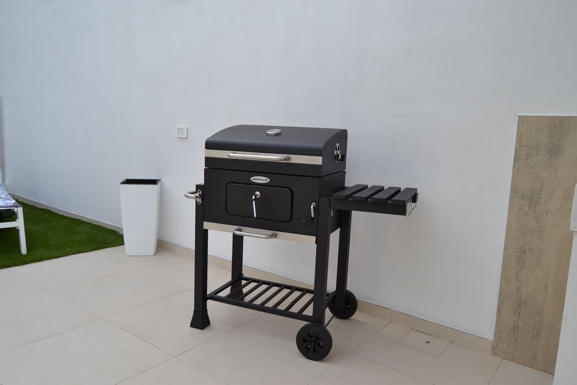 Barbecue nero da esterno con ripiano laterale e ruote, posizionato su un patio contro un muro bianco.