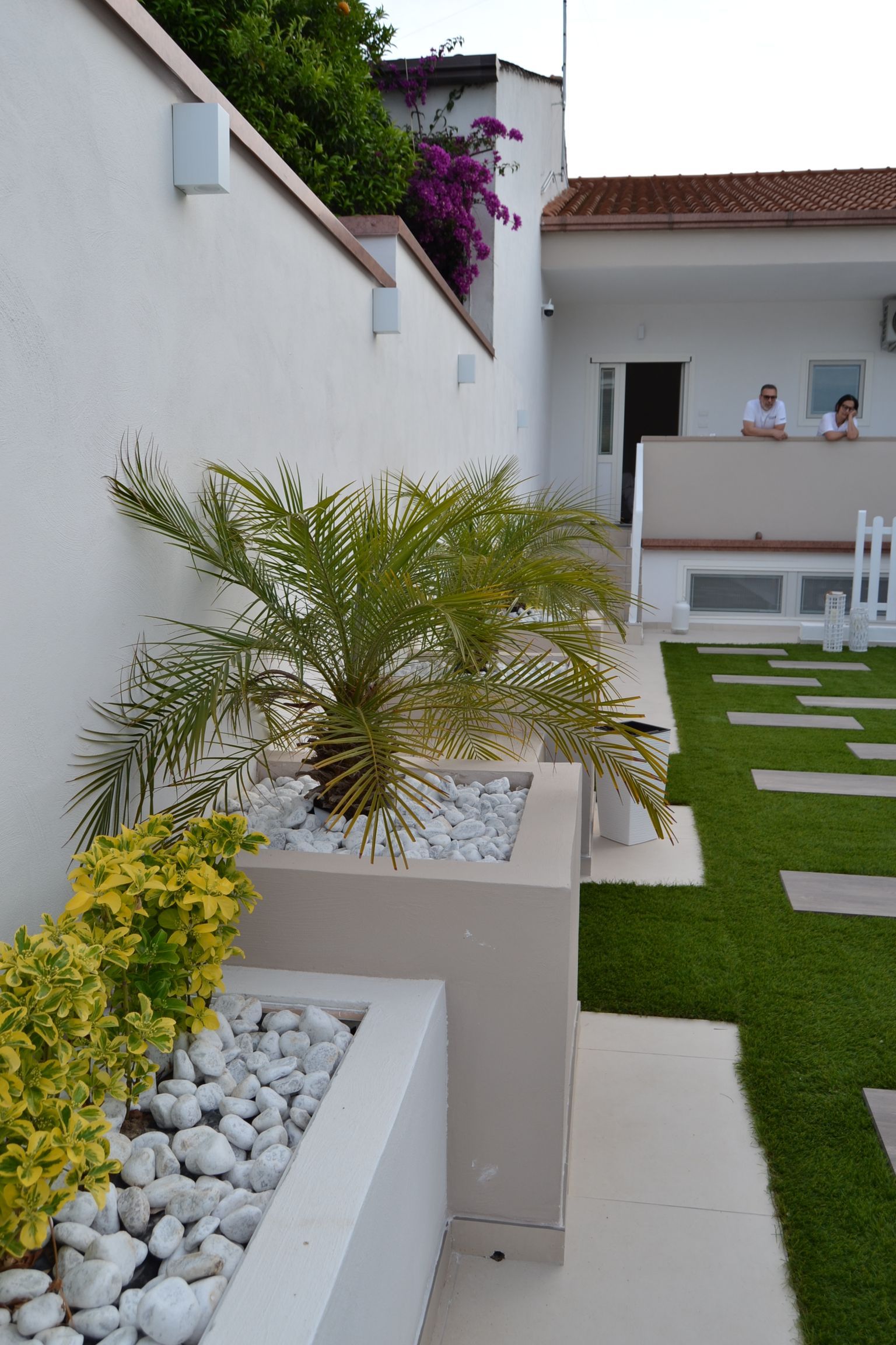 Cortile moderno con palme in vaso, muri bianchi, gradini in pietra e un balcone sullo sfondo.