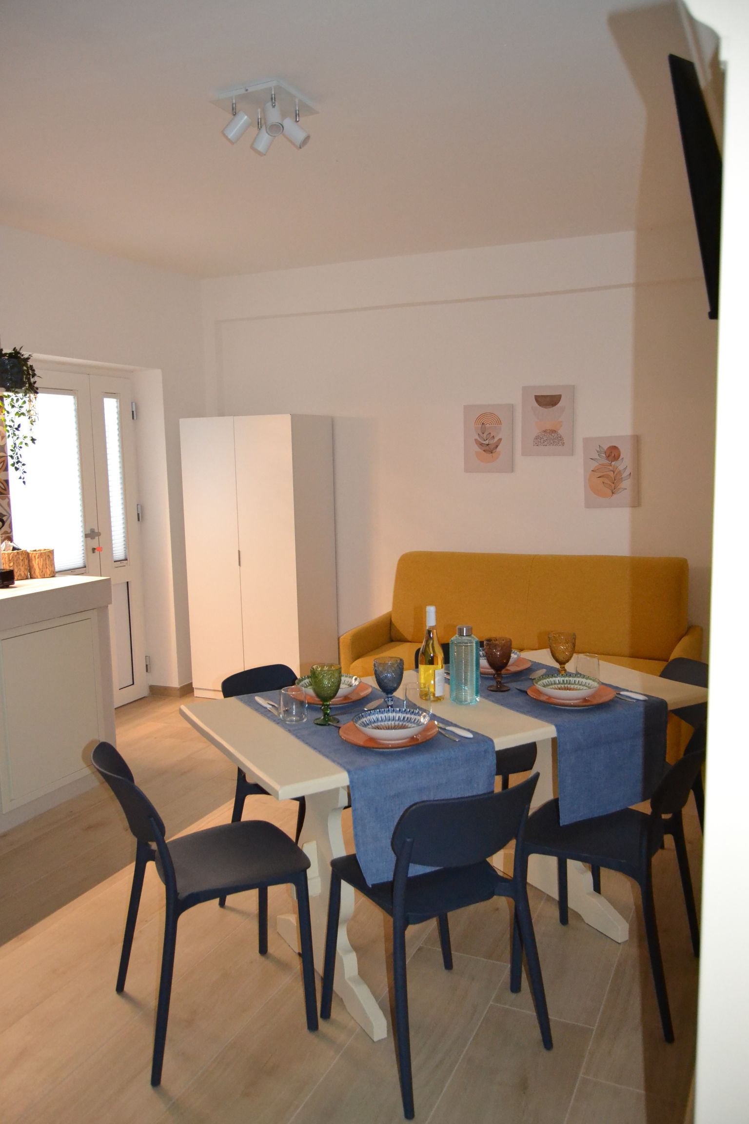Zona pranzo con tavolo ricoperto di blu, sedie nere, divano giallo e armadietti bianchi in una stanza luminosa