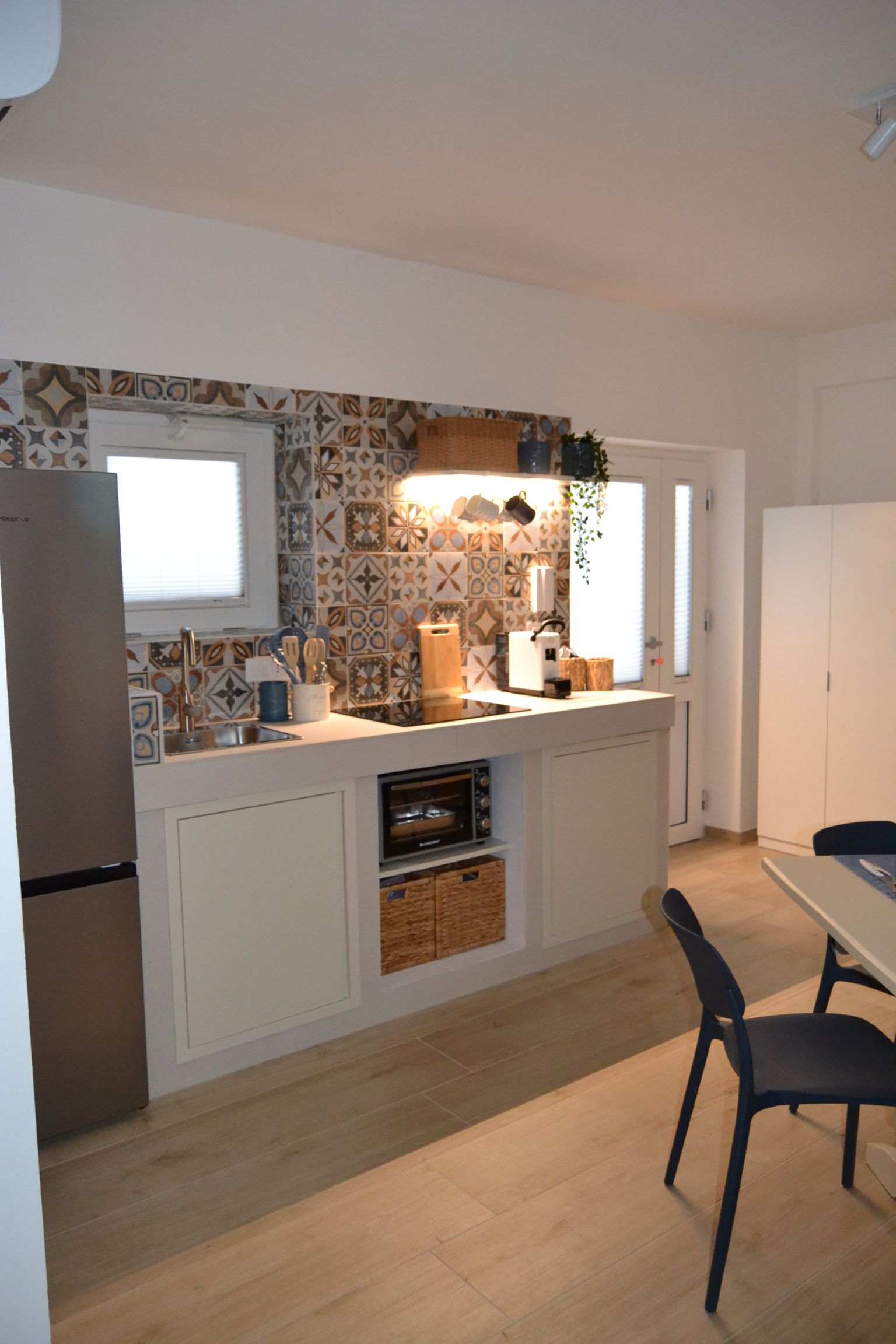 Piccola cucina con mobili bianchi, paraschizzi a mosaico e tavolo e sedie in primo piano.