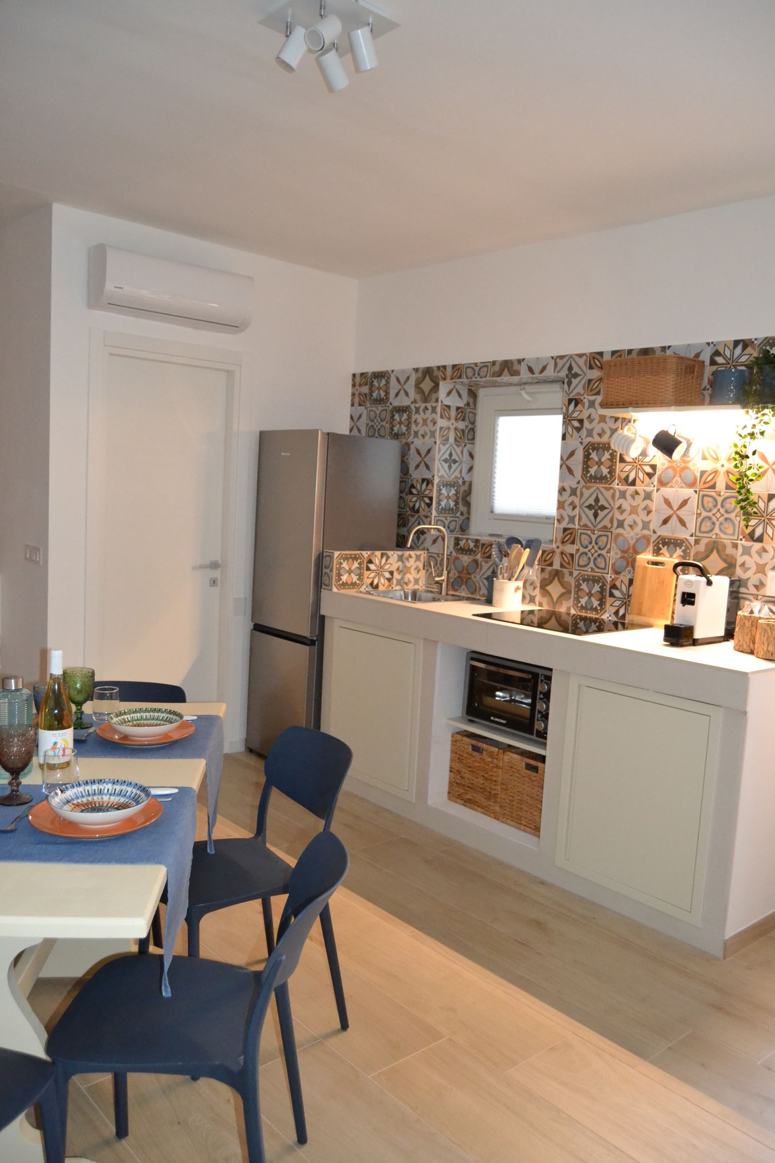 Cucina e zona pranzo compatte con sedie blu, tavolo apparecchiato, mobili bianchi e frigorifero in acciaio inox.