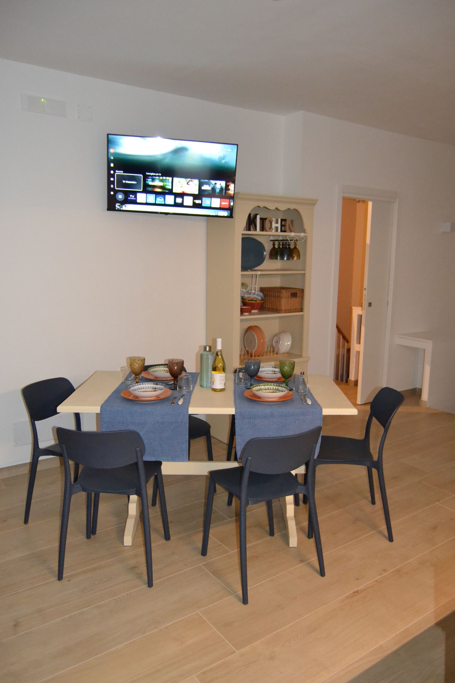 Zona pranzo con tavolo rotondo apparecchiato per un pasto, sedie, TV a parete e porta che conduce a una cucina illuminata.