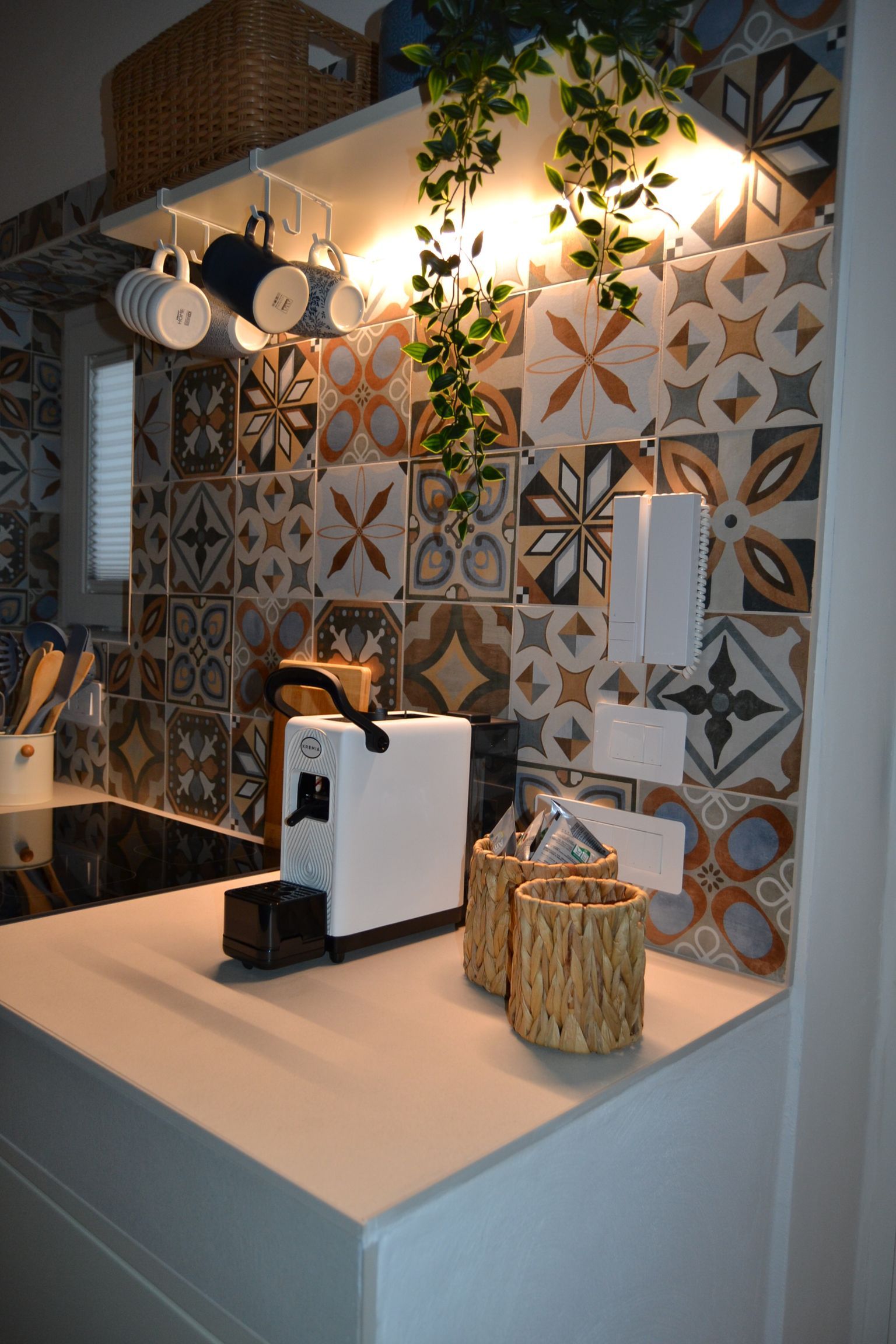 Bancone da cucina decorativo con rivestimento in piastrelle decorate, elettrodomestici bianchi e piante sospese sotto una luce calda.