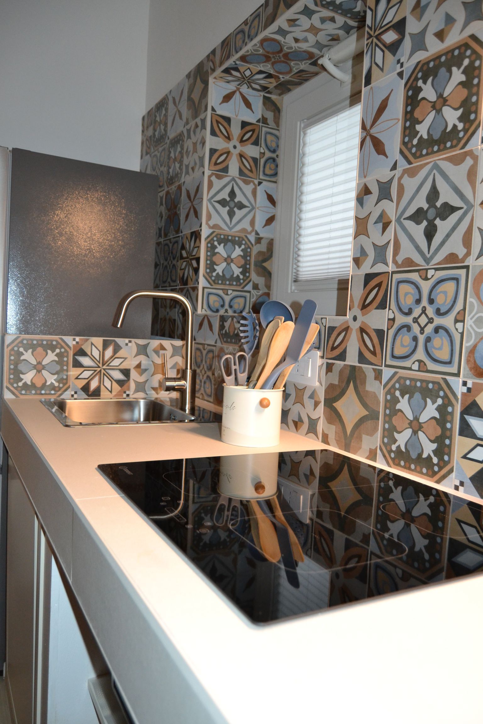 Piano di lavoro da cucina moderno con rivestimento in piastrelle decorate, piano cottura nero e portautensili vicino a una finestra.