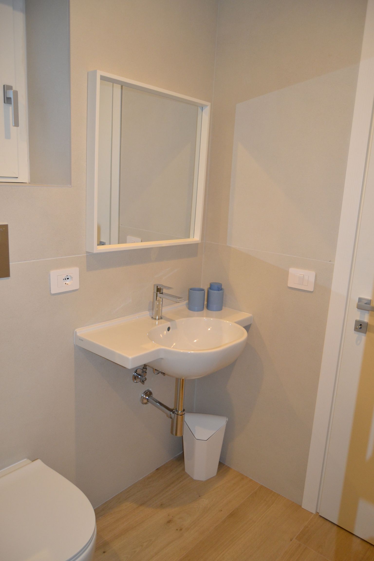Angolo bagno compatto con lavabo bianco, mobiletto con specchio, prese elettriche a muro e pavimento in legno chiaro.