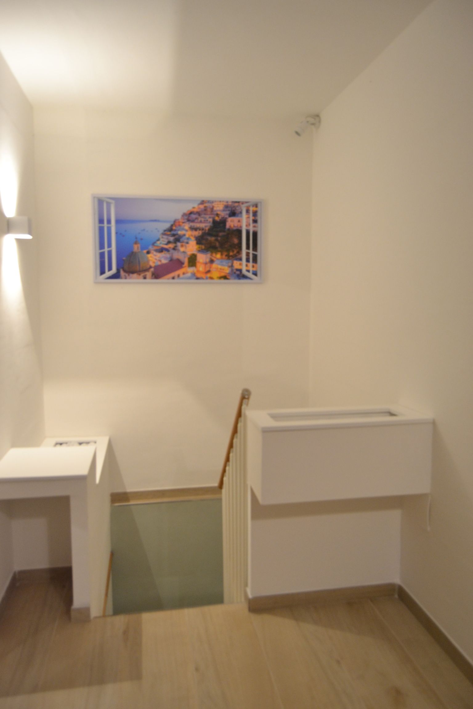 Pianerottolo delle scale con apertura sul pavimento in vetro, pareti bianche e un quadro incorniciato raffigurante un paesaggio marino.