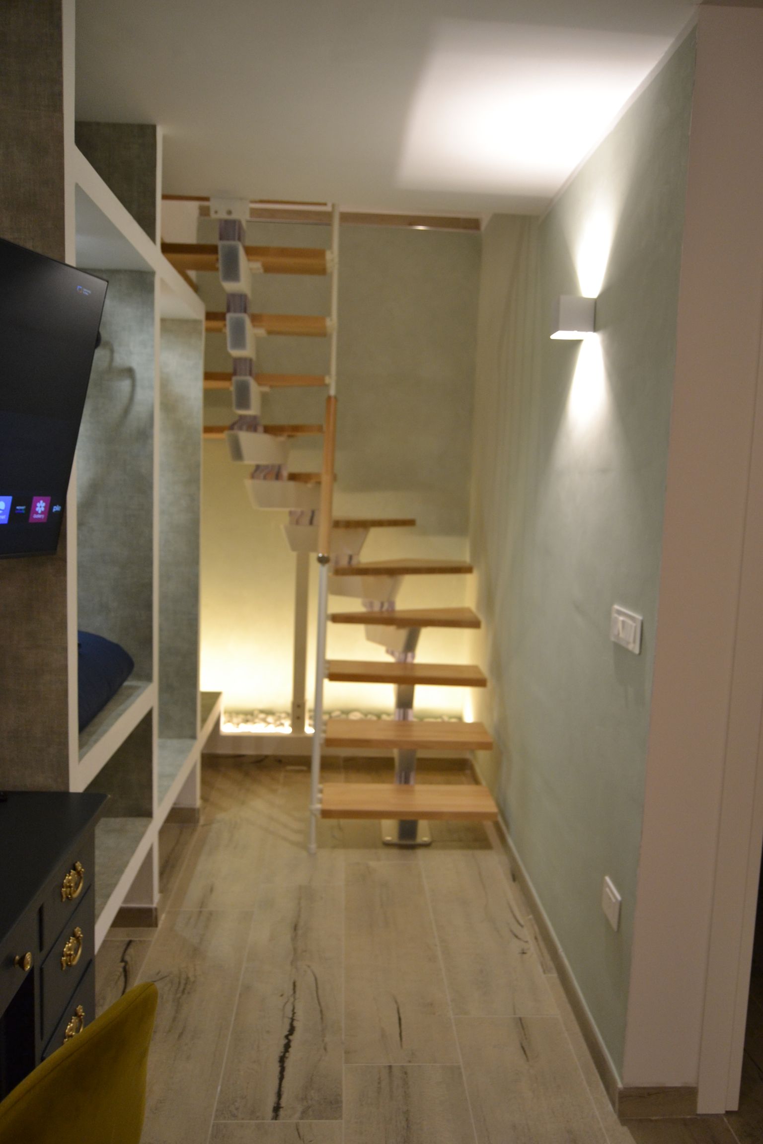 Corridoio moderno con scala in legno, pareti chiare e scaffalature a muro sotto luci calde.