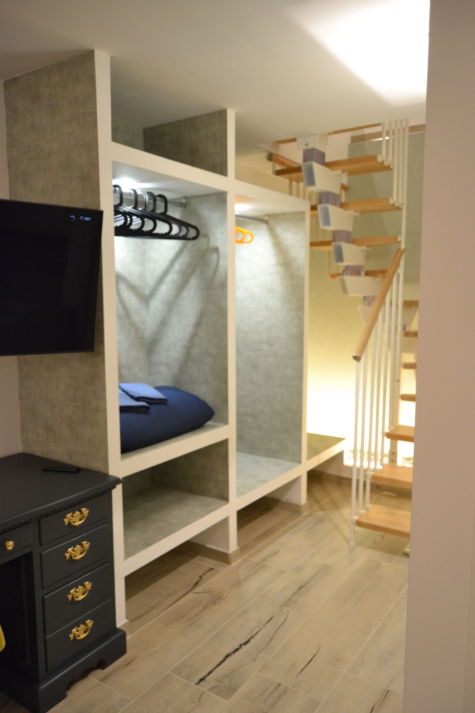 Camera da letto mansardata moderna con armadio a vista, TV, cassetti e scala in legno che conduce a un letto a soppalco.