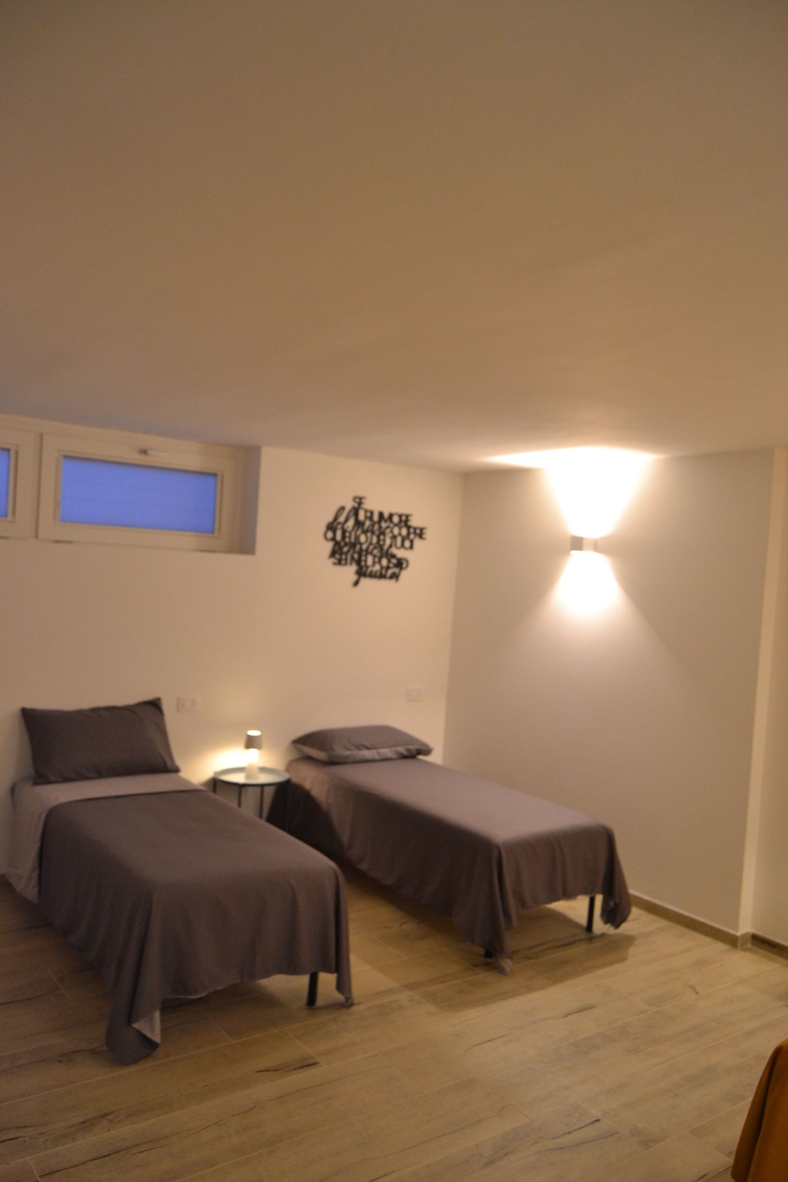 Due letti singoli in una camera da letto minimalista con pareti bianche, pavimento in legno e applique a parete.