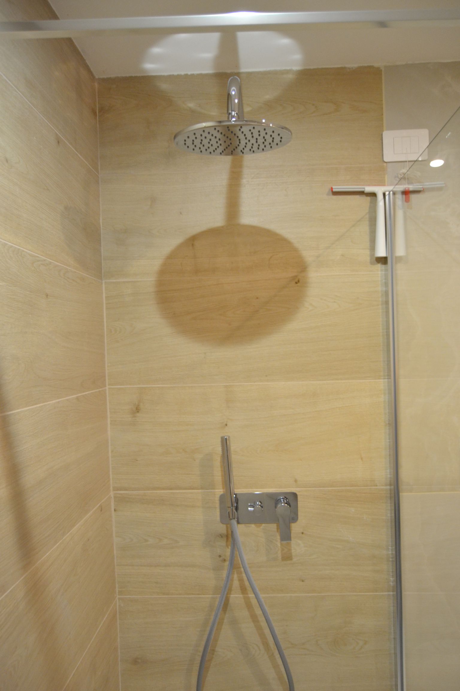 Box doccia con piastrelle beige, soffione a pioggia, doccetta a mano e maniglione di sostegno a parete.