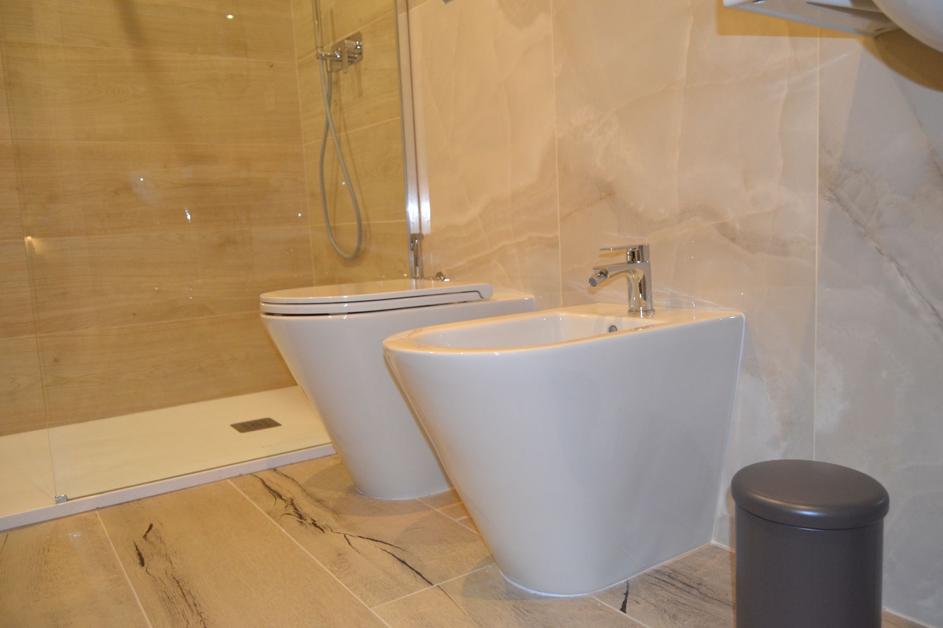 Bagno moderno con WC bianco, bidet, doccia in vetro e pareti e pavimento rivestiti con piastrelle beige.