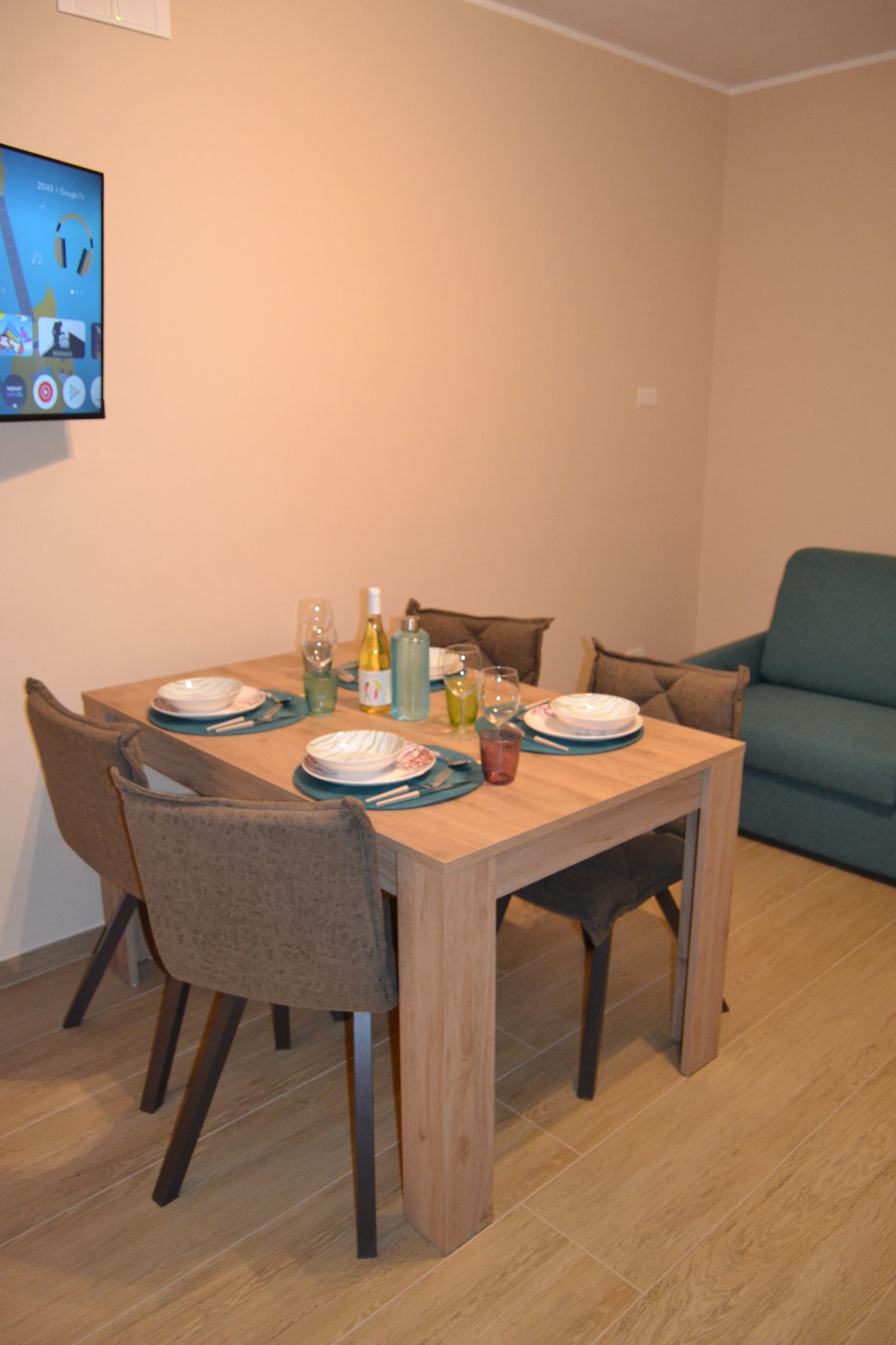 Tavolo da pranzo apparecchiato con piatti in un soggiorno beige accanto a un divano blu e a una TV a parete.