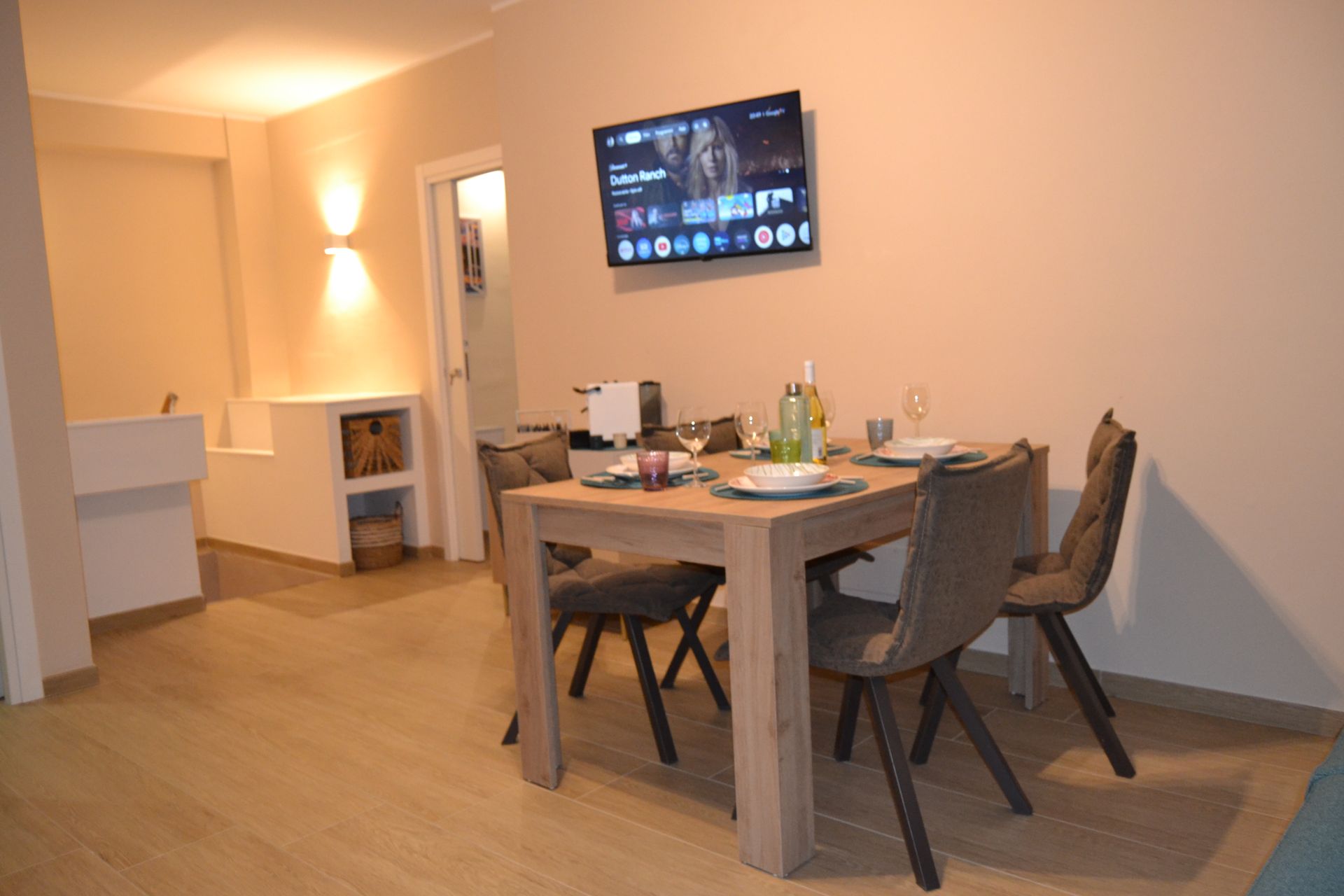 Zona pranzo moderna con tavolo in legno, quattro sedie, TV a parete e illuminazione calda.