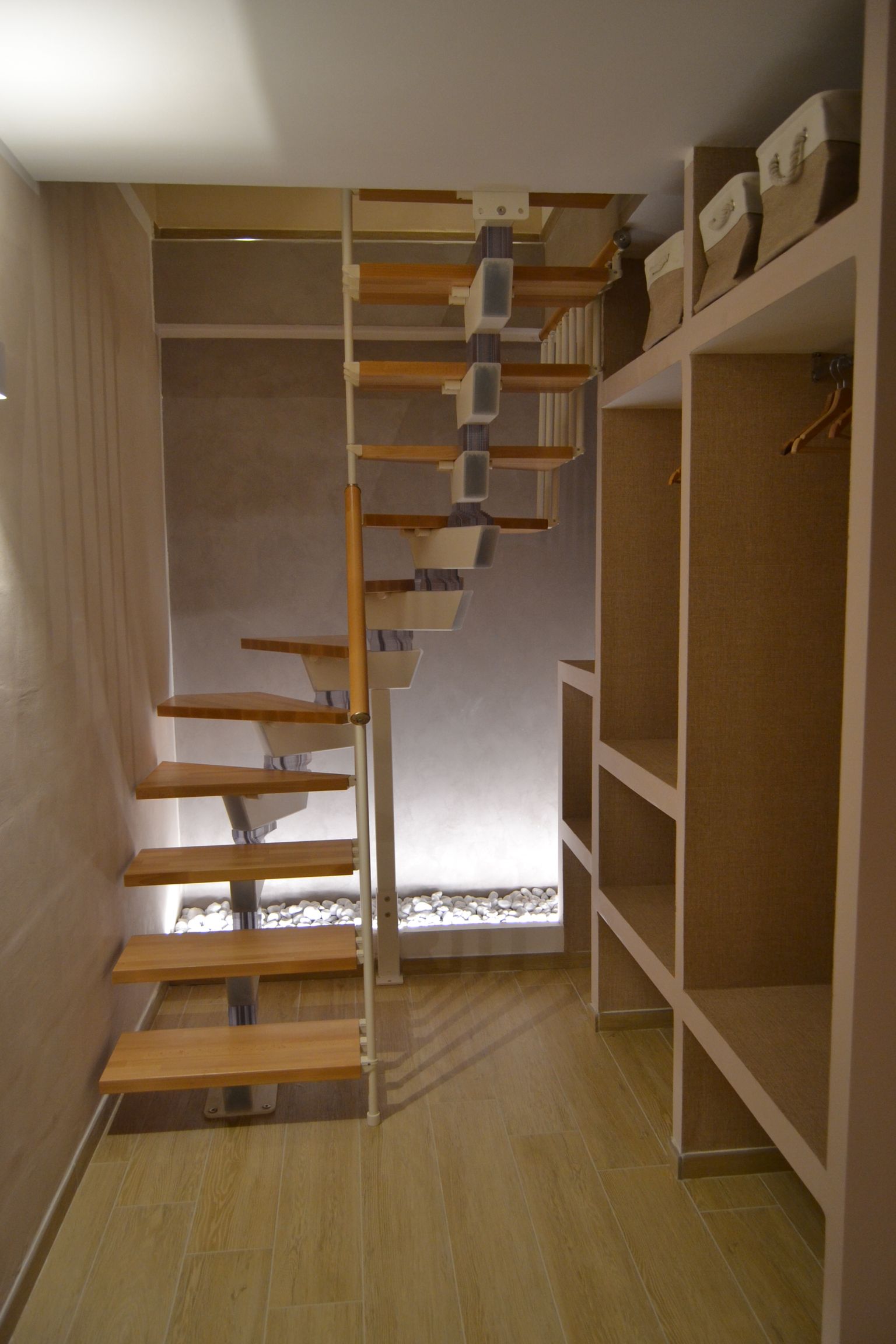 Stretta scala in legno all'interno di una stanza simile a un ripostiglio con scaffalature e un'apertura sul soppalco.