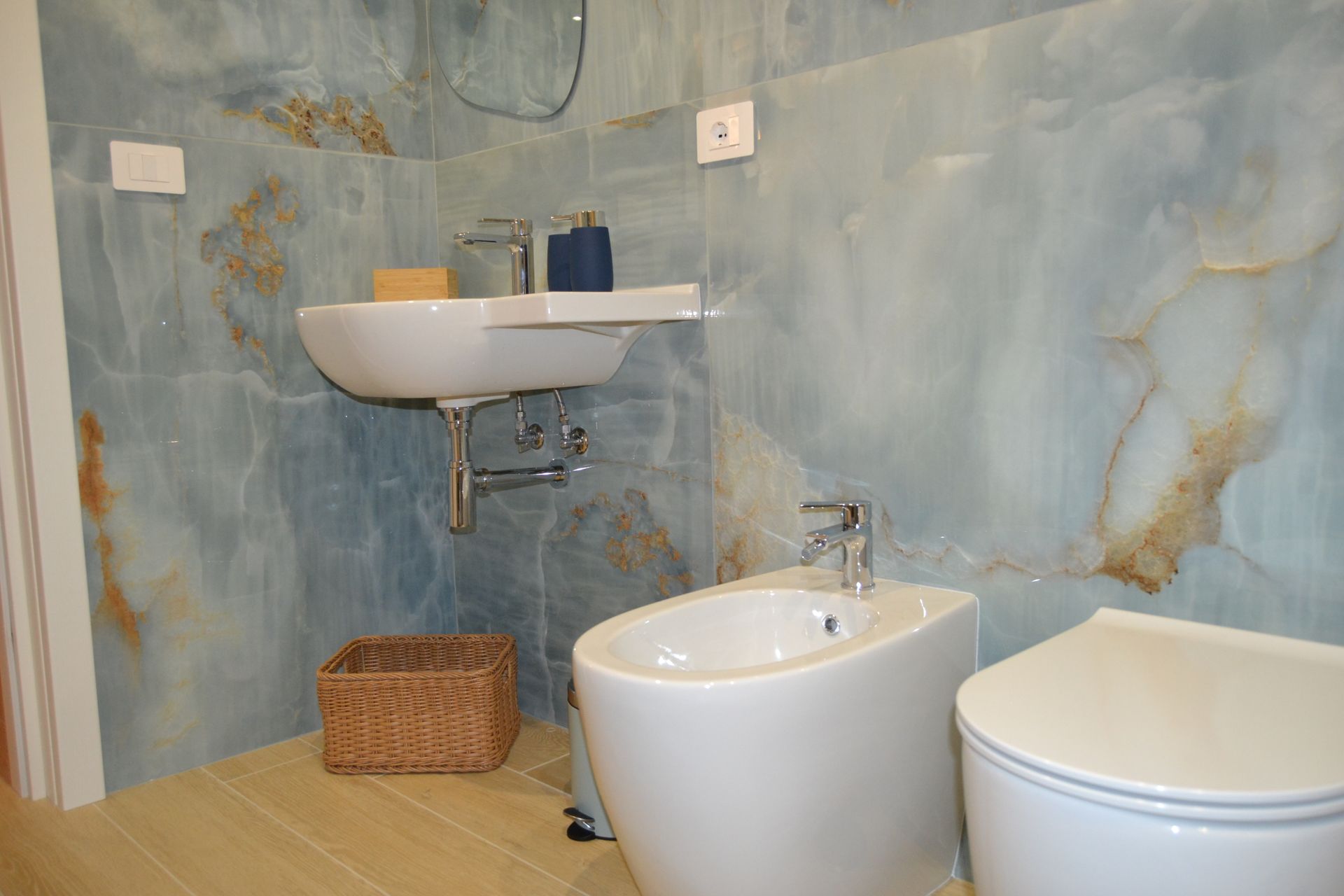 Bagno moderno con pareti in marmo blu, lavabo bianco, bidet, WC e cesto di vimini sul pavimento.