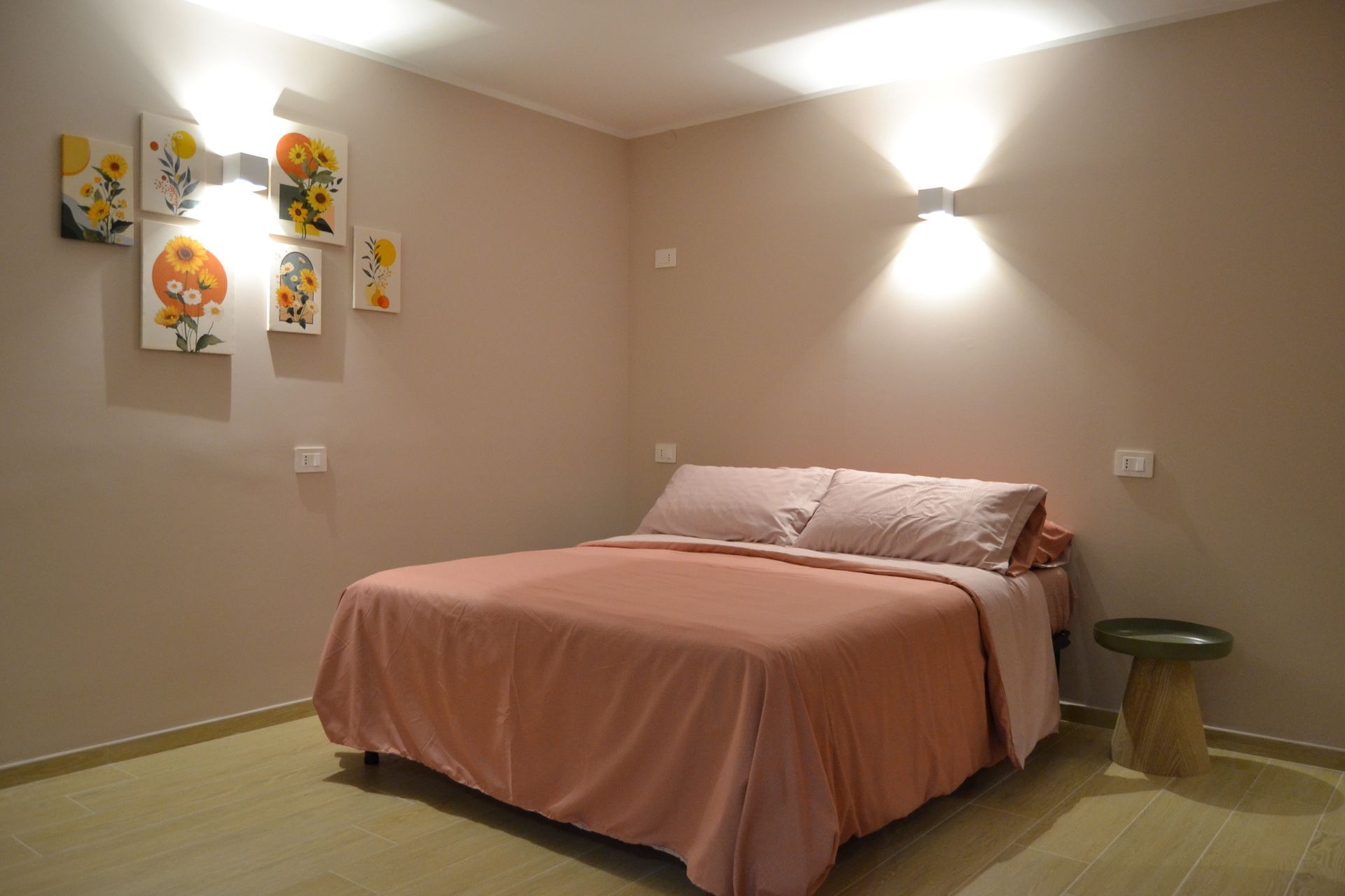 Camera da letto minimalista con copriletto rosa, quadri alle pareti e pareti beige illuminate da due applique.