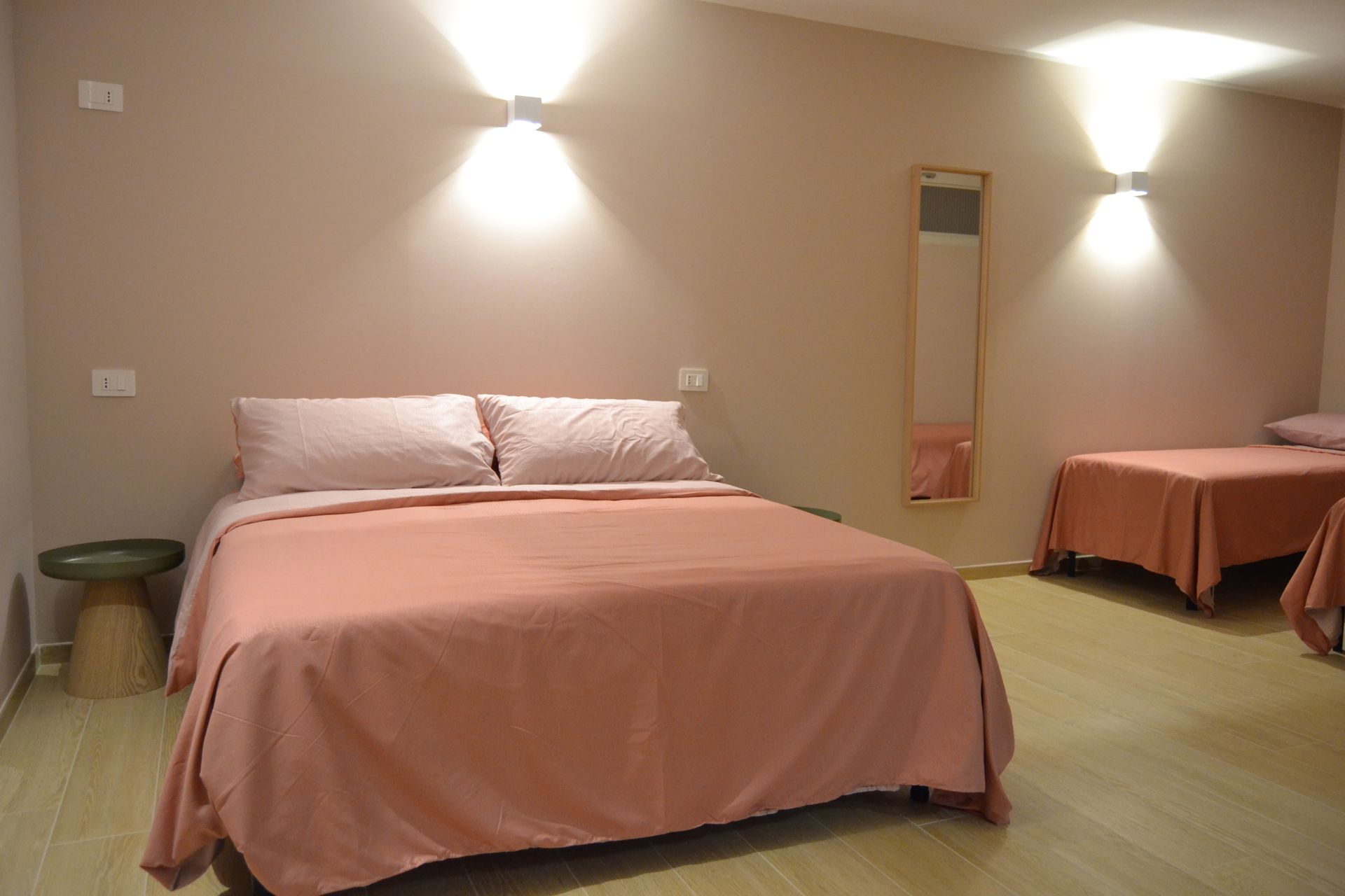 Camera da letto con copriletto rosa, letto singolo, applique e un piccolo comodino in una stanza beige.