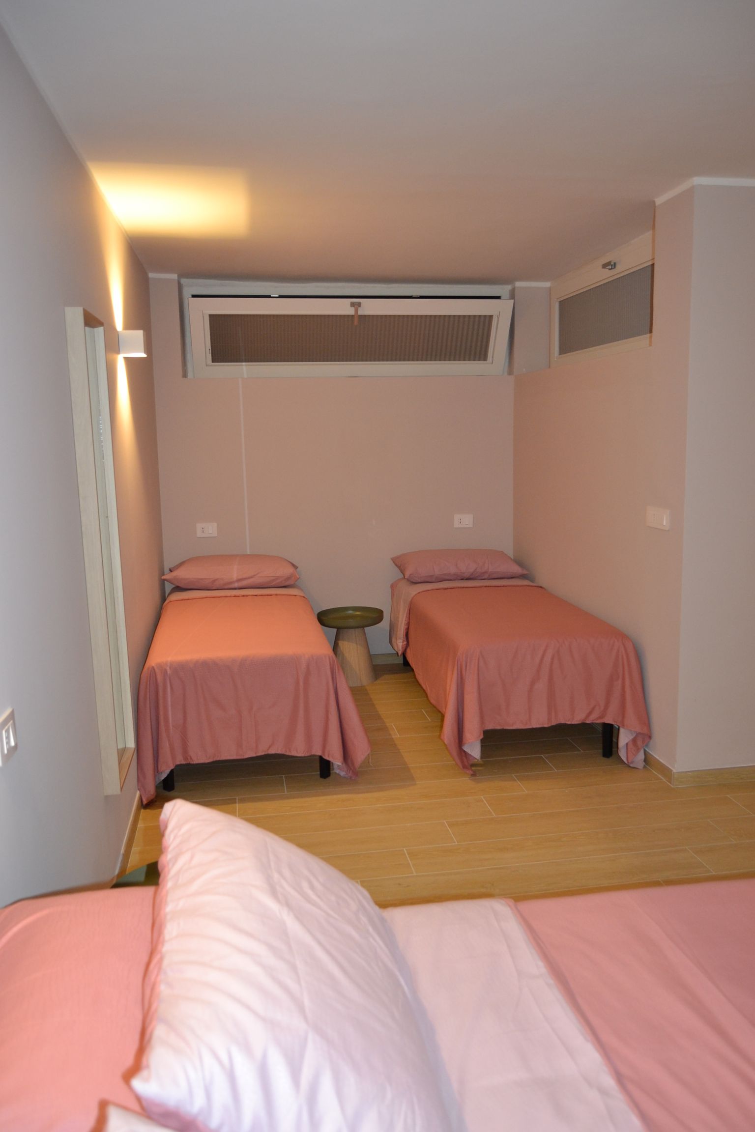 Piccola camera d'albergo con tre letti singoli rosa e un condizionatore a finestra.