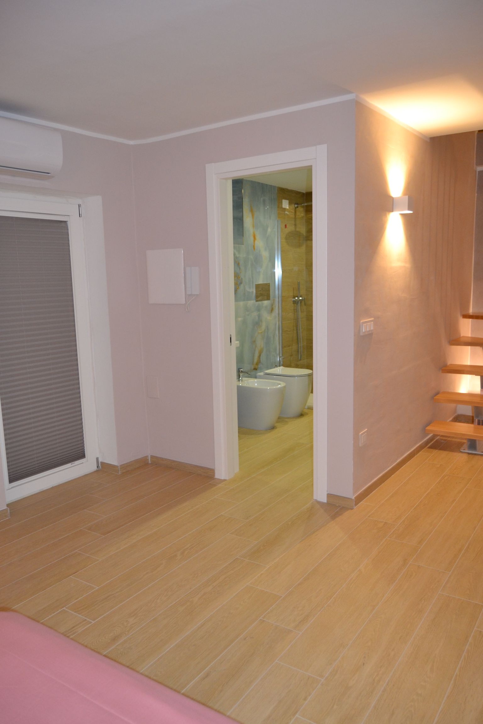 Camera da letto vuota con pavimento in legno chiaro, porta del bagno aperta e scale illuminate sulla destra.
