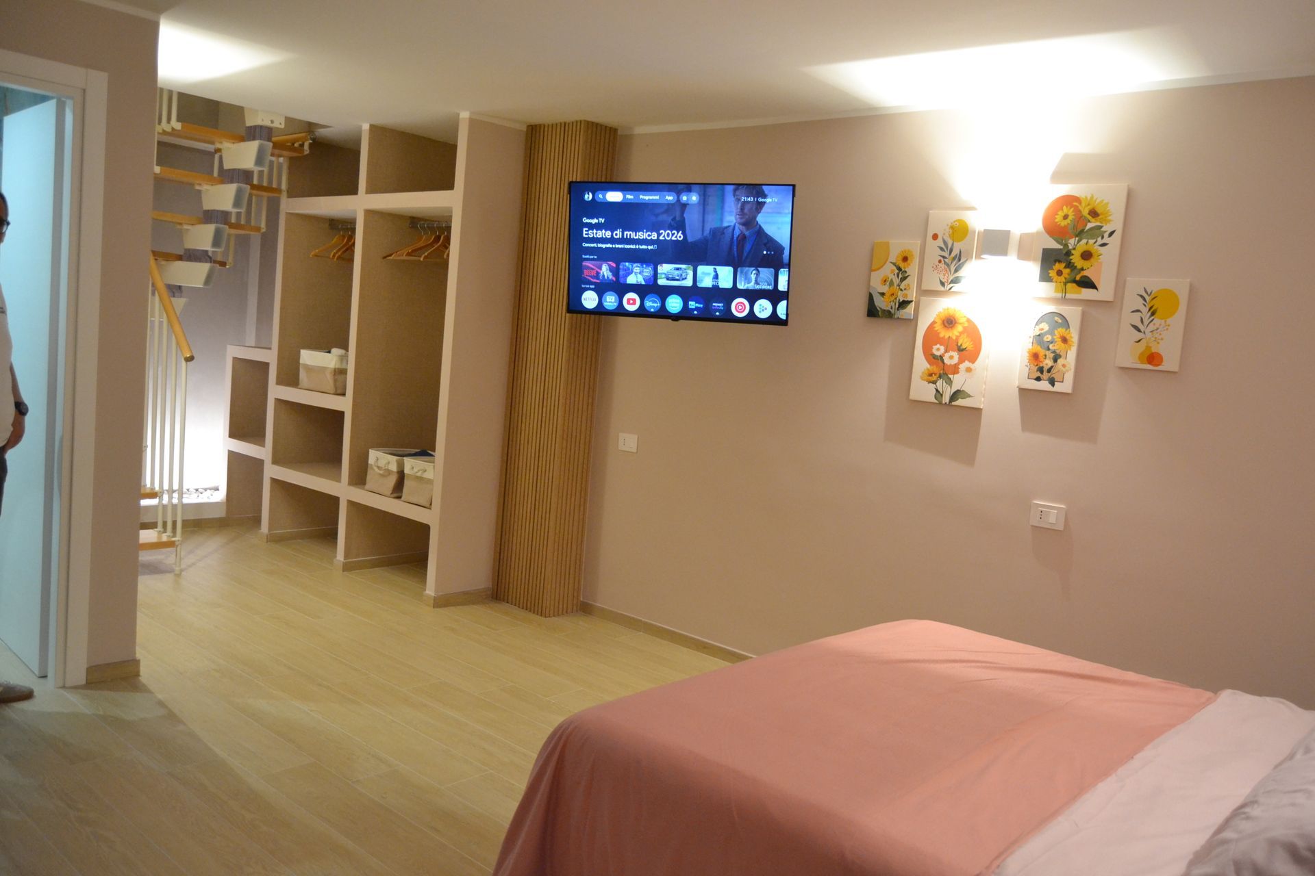 Camera da letto moderna con copriletto rosa, TV a parete, mensole a vista e decorazioni floreali alle pareti.