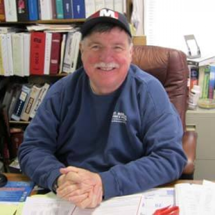 A man wearing a hat with the letter m on it is sitting at a desk.
