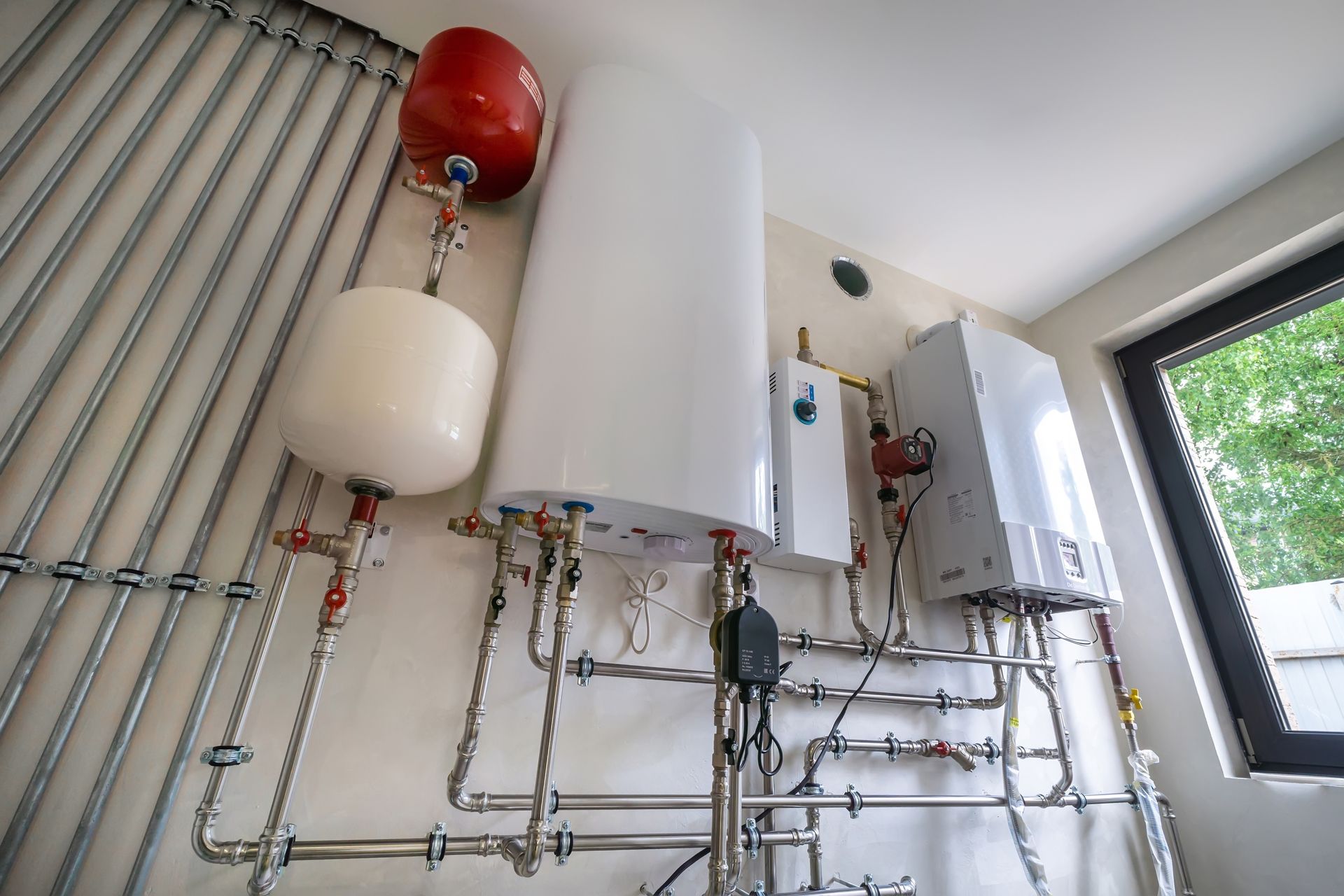 Plumbing system with white and red tanks, pipes, and a window in a white-walled room.