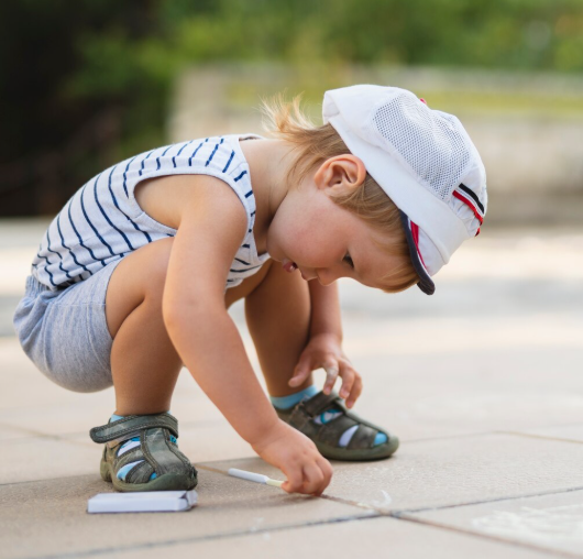 Un niño con camiseta y gorra a rayas se agacha para dibujar con tiza en el pavimento.