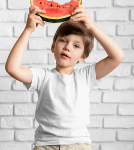 Niño que sostiene una rodaja de sandía sobre su cabeza, de pie frente a una pared de ladrillo blanco.