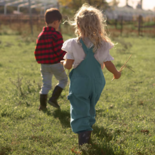 Dos niños caminan por un campo de hierba. Uno lleva un mono azul, el otro una camisa de cuadros roja.