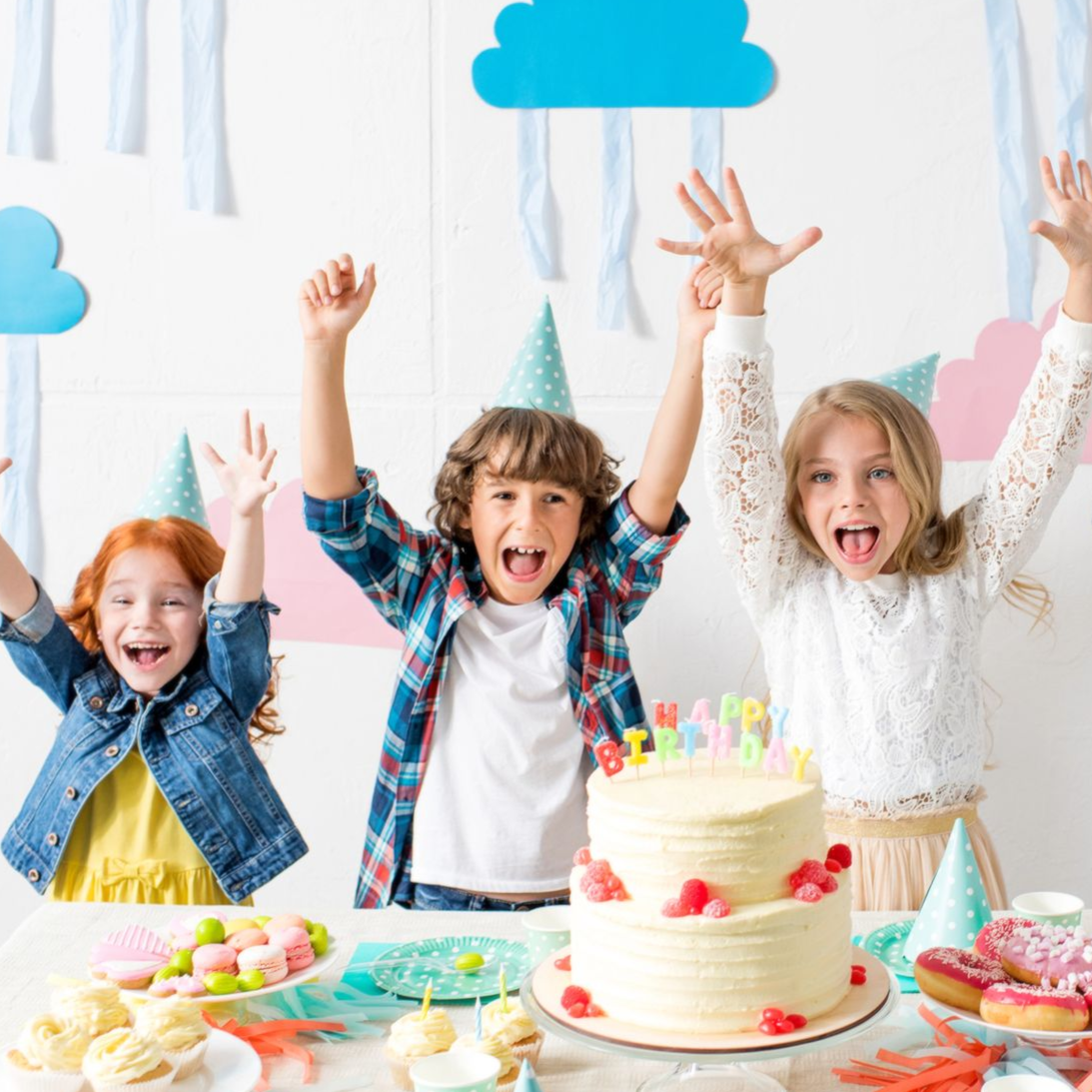 Tres niños emocionados en una fiesta de cumpleaños, con los brazos en alto, junto a un pastel con velas encendidas.