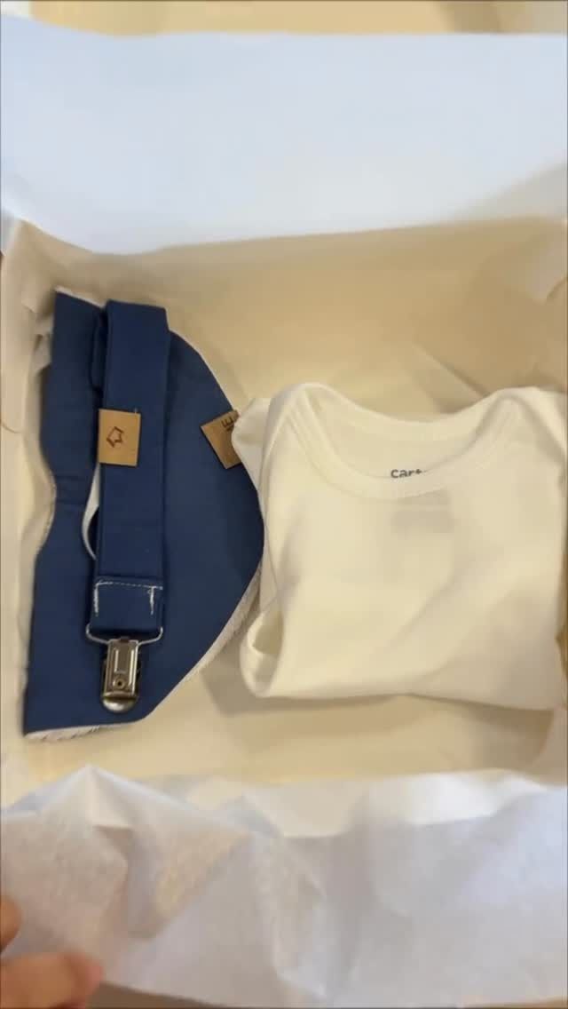 Una caja blanca contiene un babero y tirantes azules, y un mono blanco.