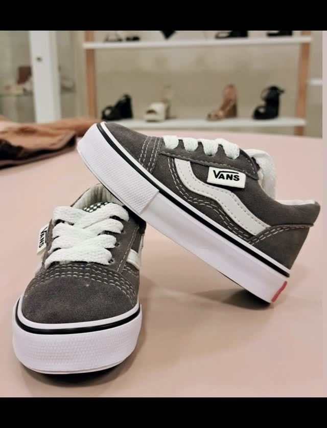 Zapatillas Vans grises y blancas sobre una superficie rosa con una estantería de tienda al fondo.